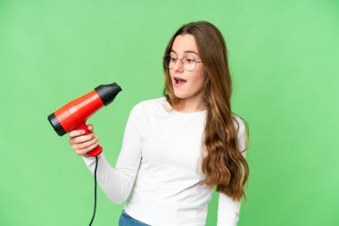 Teenager girl holding a hairdryer over isolated chroma key background with surprise and shocked facial expression