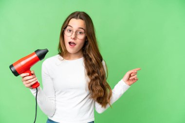 Teenager girl holding a hairdryer over isolated chroma key background surprised and pointing side