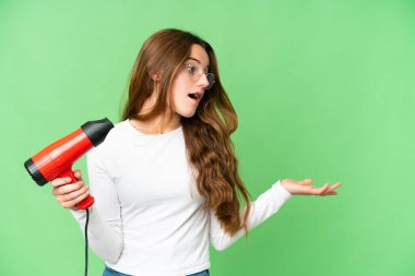 Teenager girl holding a hairdryer over isolated chroma key background with surprise facial expression
