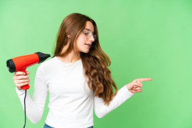 Teenager girl holding a hairdryer over isolated chroma key background pointing to the side to present a product
