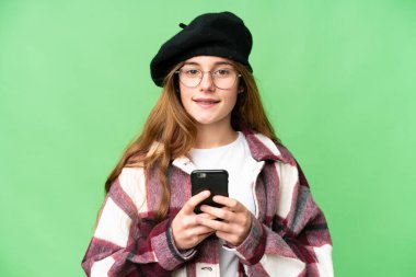 Teenager girl over isolated chroma key background looking at the camera and smiling while using the mobile