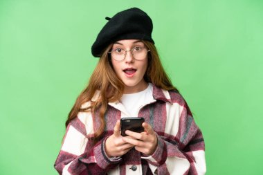 Teenager girl over isolated chroma key background looking at the camera while using the mobile with surprised expression