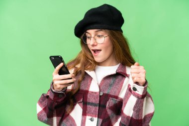 Teenager girl over isolated chroma key background using mobile phone and doing victory gesture