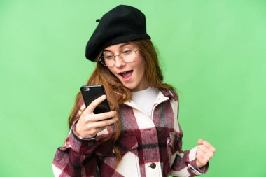 Teenager girl over isolated chroma key background using mobile phone and doing victory gesture