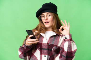 Teenager girl over isolated chroma key background using mobile phone and doing OK sign