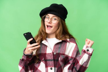 Teenager girl over isolated chroma key background using mobile phone and doing victory gesture