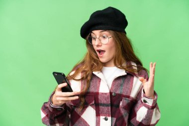 Teenager girl over isolated chroma key background looking at the camera while using the mobile with surprised expression