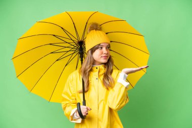 Yağmura dayanıklı ceketi ve şemsiyesi olan genç bir kız izole edilmiş krom anahtar arkaplanı üzerinde. Davet etmek için ellerini yana uzatıyor.