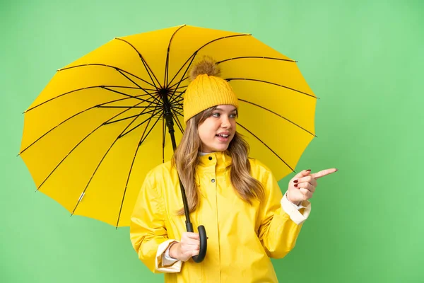 年轻姑娘穿着防雨外套和雨伞 罩在孤立的彩色关键背景上 手指指向侧面 并展示了一种产品 — 图库照片