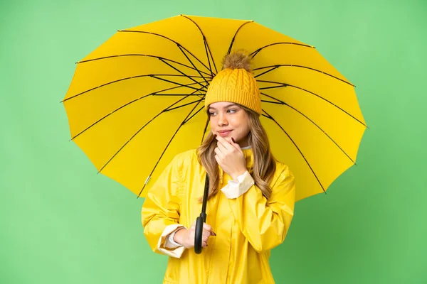 若いです女の子とともにレインプルーフコートと傘上の隔離されたクロマキー背景見ますザ側と笑顔 — ストック写真