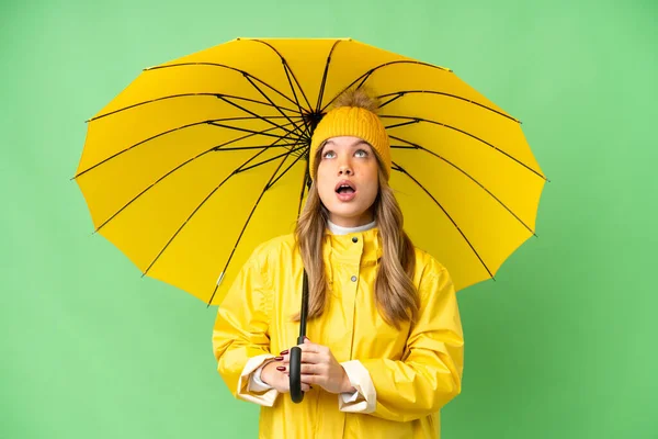 穿着雨衣 披着雨伞的小女孩 在孤零零的彩色关键背景上抬起头 带着惊讶的表情 — 图库照片
