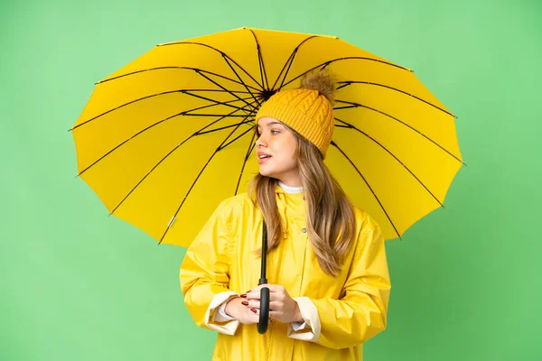 年轻姑娘穿着防雨外套和雨伞 披着孤零零的彩色关键背景 朝旁边看去 面带微笑 — 图库照片
