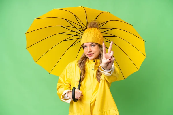 穿着雨衣 披着雨伞的小女孩 在孤零零的彩色关键背景上微笑着 展现出胜利的迹象 — 图库照片