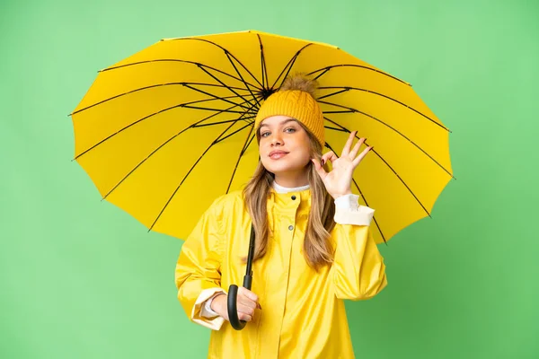 若いです女の子とともにレインプルーフコートと傘上の隔離されたクロマキー背景ショーOk Signとともに指 — ストック写真