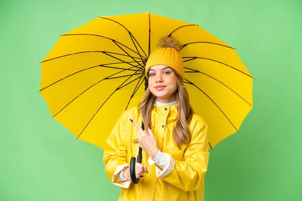 若いです女の子とともにレインプルーフコートと傘上の隔離されたクロマキー背景指す側に製品を提示 — ストック写真