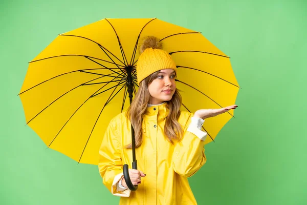 年轻姑娘穿着防雨外套 用雨伞遮挡着孤立的彩色关键背景 向旁边伸出手来 邀请她来 — 图库照片