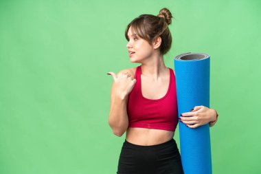 Genç sporcu kız yoga derslerine giderken izole edilmiş krom anahtar arkaplanının üzerinde bir minder tutuyor ve bir ürünü sunmak için yan tarafı işaret ediyor.