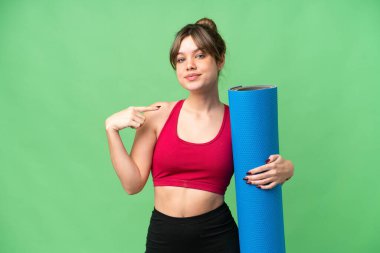 Genç sporcu kız yoga derslerine giderken izole edilmiş krom anahtar arkaplanının üzerinde paspas tutuyor ve baş parmak hareketi yapıyor.