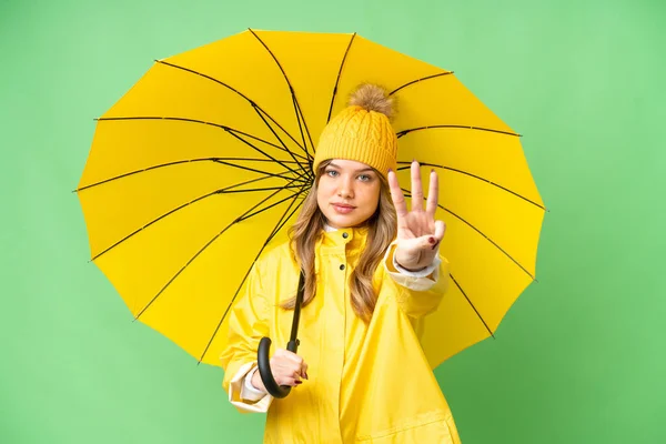 年轻姑娘穿着雨衣 带着雨伞 在孤立的彩色关键背景上快乐地数着三个手指 — 图库照片