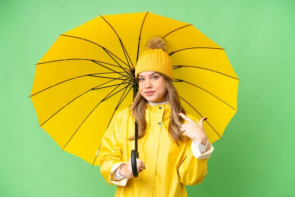 穿着防雨外套 披着雨伞 戴在孤立的彩色关键背景上的小女孩举起了一个大拇指 — 图库照片