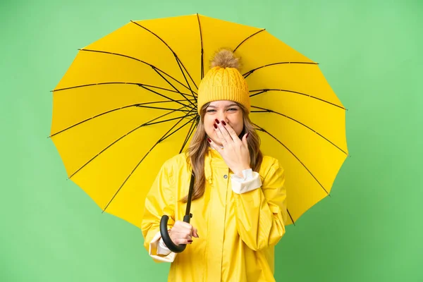 年轻姑娘穿着雨衣 披着雨伞 披着孤零零的彩色关键背景 幸福地微笑着 用手捂着嘴 — 图库照片