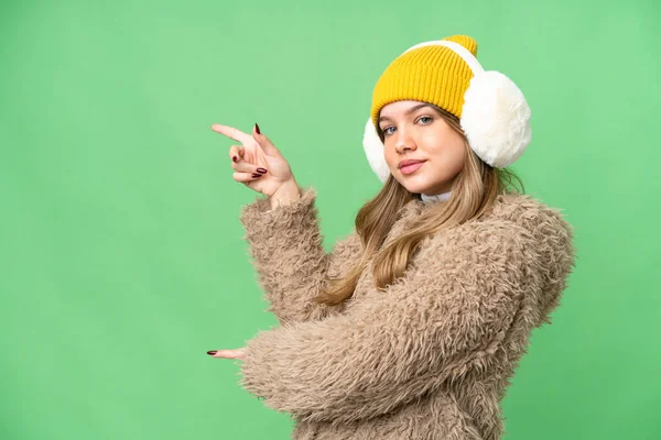 年轻姑娘披着冬衣 罩在孤立的彩色关键背景上 指指点点旁边 展示自己的产品 — 图库照片