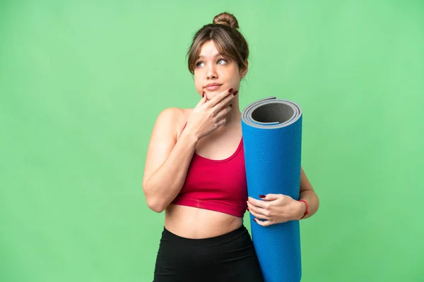 年轻的女运动员一边去上瑜伽课 一边拿着垫子盖着孤立的彩色关键背景 心里充满了疑虑 — 图库照片
