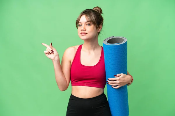 年轻的女运动员一边去上瑜伽课 一边拿着垫子 把手指指向旁边 用孤立的彩色键做背景 — 图库照片