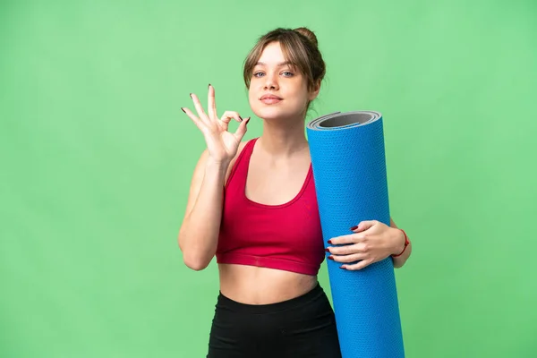 年轻的女运动员一边去上瑜伽课 一边拿着垫子 用手指在孤立的色键背景上显示出好的手势 — 图库照片