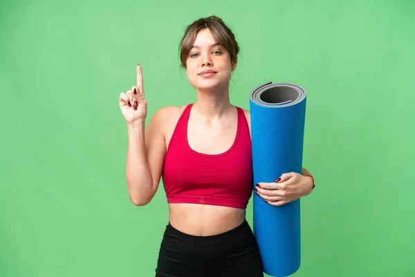 参加瑜伽课的年轻女运动员拿着一个垫子 盖住一个孤立的彩色键背景 展示并举起一个手指 表示她是最好的 — 图库照片