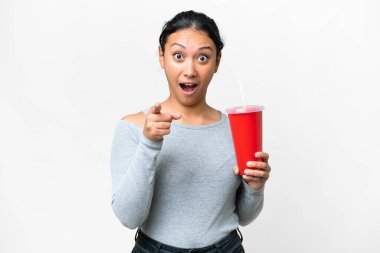 Young Uruguayan woman holding soda over isolated white background surprised and pointing front