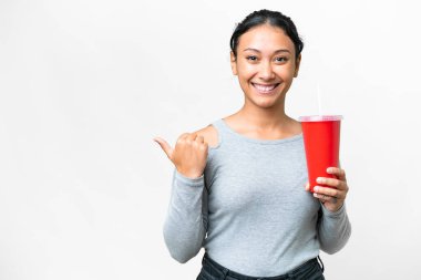Young Uruguayan woman holding soda over isolated white background pointing to the side to present a product