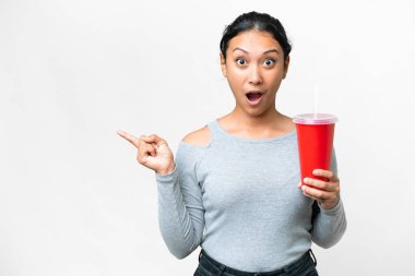 Genç Uruguaylı kadın izole edilmiş beyaz arka plan üzerinde soda tutuyor. Şaşırmış ve işaret ediyor.
