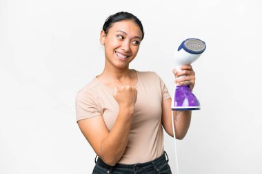 Young Uruguayan woman holding a vertical steam iron over isolated white background celebrating a victory