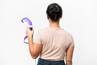 Young Uruguayan woman holding a vertical steam iron over isolated white background in back position