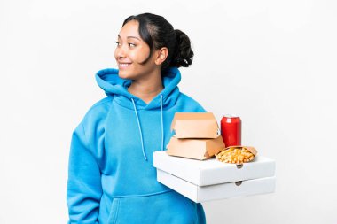 Young Uruguayan woman holding pizzas and burgers over isolated white background looking side