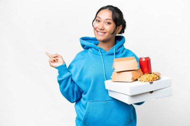 Young Uruguayan woman holding pizzas and burgers over isolated white background pointing finger to the side