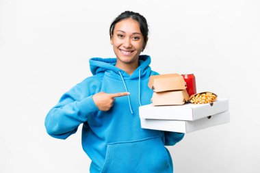 Young Uruguayan woman holding pizzas and burgers over isolated white background pointing to the side to present a product