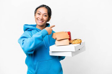 Young Uruguayan woman holding pizzas and burgers over isolated white background pointing back