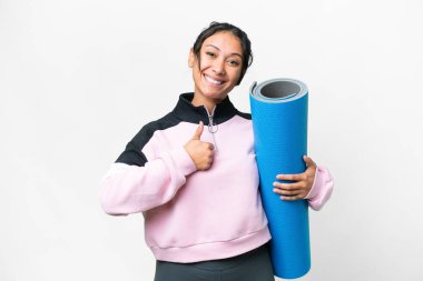 Genç sporcu kadın yoga derslerine giderken izole edilmiş beyaz arka plan üzerinde bir minder tutuyor ve baş parmak hareketi yapıyor.