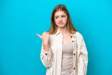 Teenager Russian girl isolated on blue background unhappy and pointing to the side