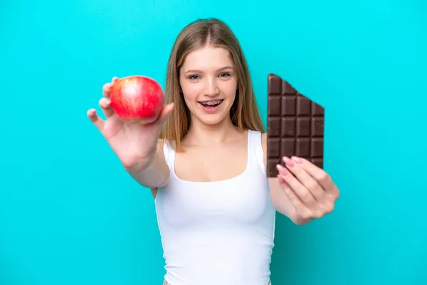 十代のロシア人の女の子は青の背景に隔離されました一方でチョコレートタブレットともう一方でリンゴを取ります — ストック写真