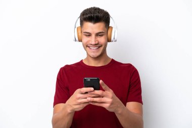 Beyaz arka planda izole edilmiş genç beyaz bir adam müzik dinliyor ve cep telefonu arıyor.