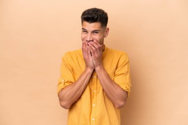 Beyazlı genç bir adam bej arka planda izole edilmiş mutlu ve gülümseyen elleriyle ağzını kapatıyor.