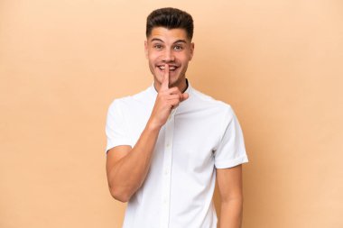 Beyazlı genç adam bej arka planda izole edilmiş. Parmağını ağzına koyduğunda sessizlik işareti gösteriyor.