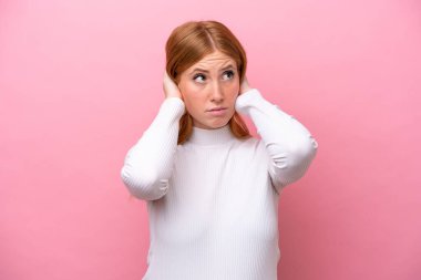 Genç kızıl saçlı kadın pembe arka planda izole edilmiş hayal kırıklığına uğramış ve kulaklarını kapatmış.