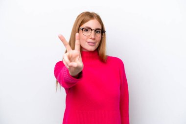 Beyaz arka planda izole edilmiş genç kızıl saçlı kadın gülümsüyor ve zafer işareti gösteriyor.