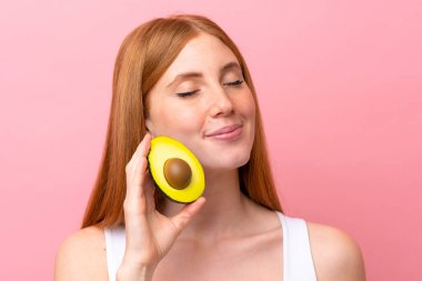 Pembe arka planda izole edilmiş genç kızıl saçlı kadın elinde bir avokado tutuyor. Portreyi kapat