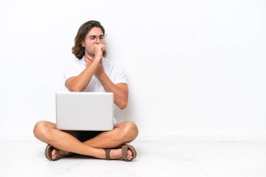 Yerde dizüstü bilgisayarı olan genç ve yakışıklı bir adam beyaz arka planda izole bir şekilde oturuyor ve öksürüp kendini kötü hissediyor.