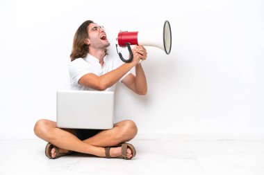 Young handsome man with a laptop sitting on the floor isolated on white background shouting through a megaphone